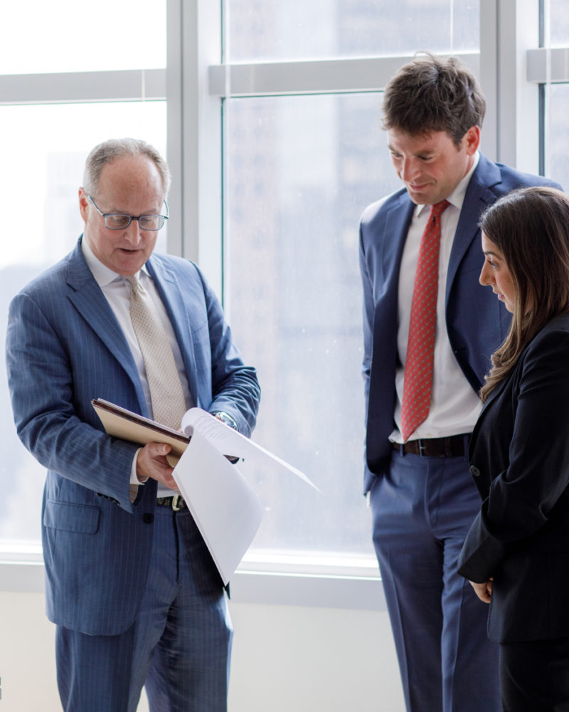 Standing in front of a glass wall, an older Cohen Clair professional shows two younger colleagues some paperwork.